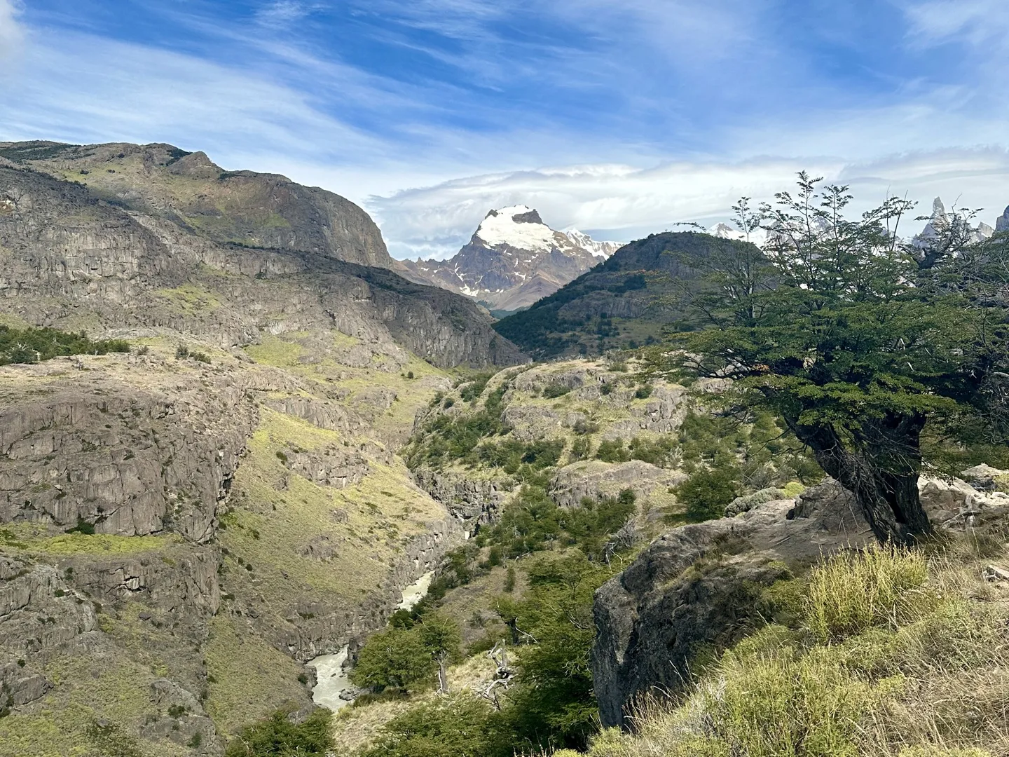 A river cutting through a rocky gorge below the trail, with a snow-capped peak on the skyline