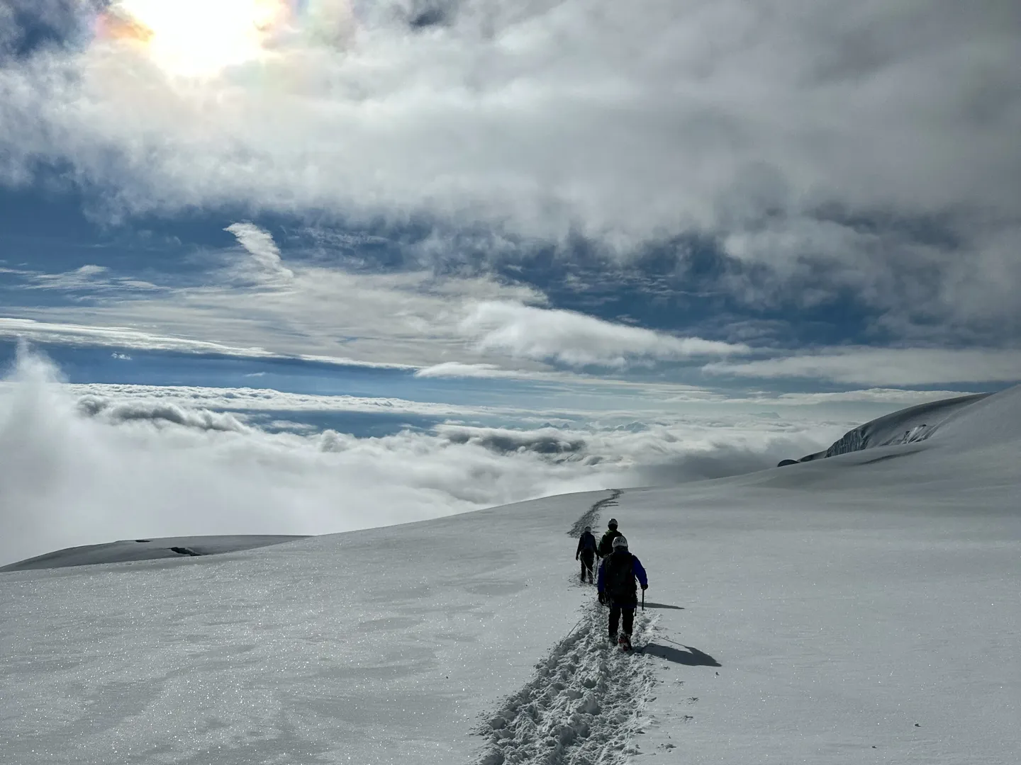 Climbers on the snow with the sun low behind cloud, a sea of cloud filling the valley below