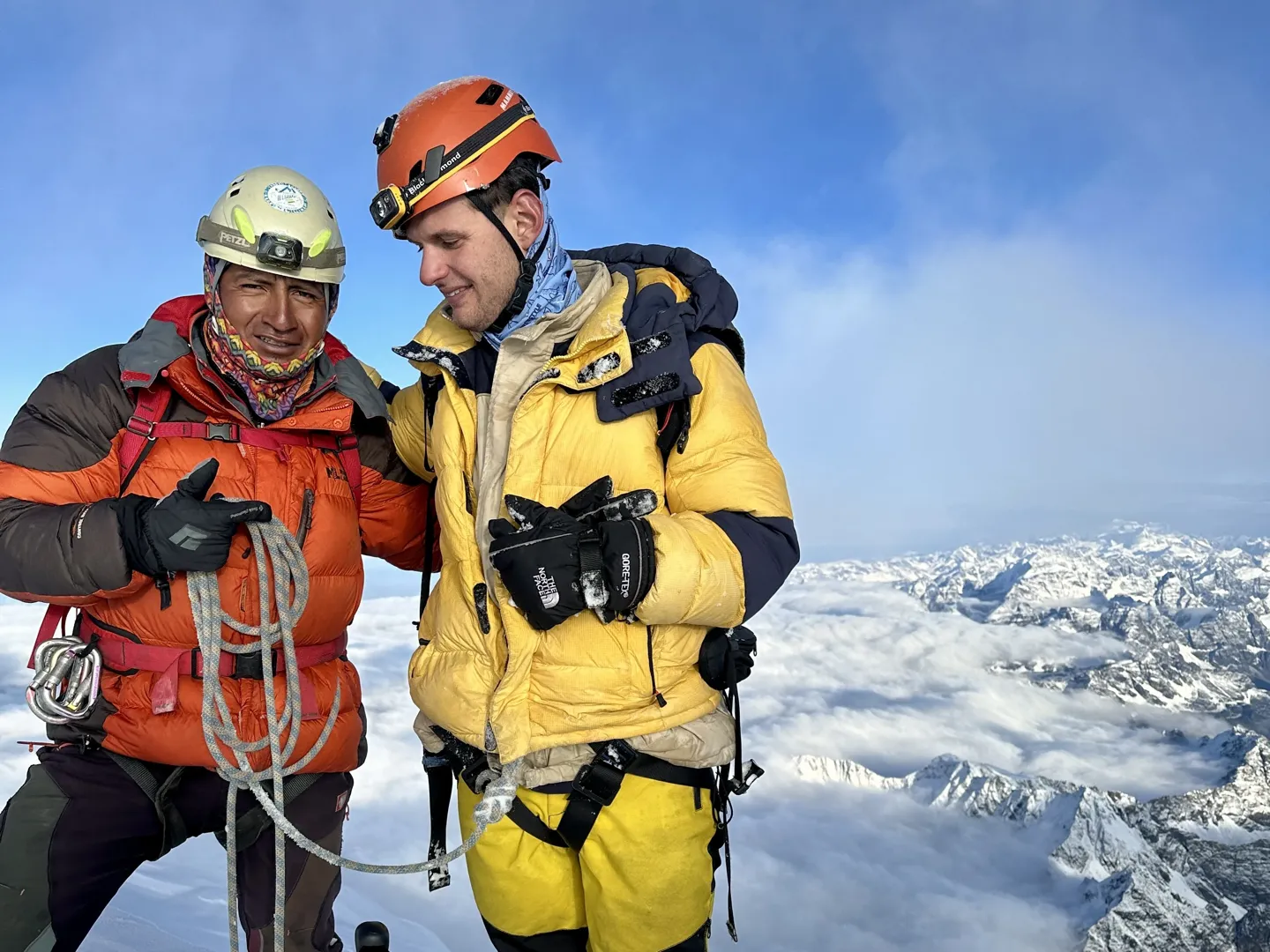 Two climbers in down jackets and helmets at the Huayna Potosi summit, cloud and peaks behind