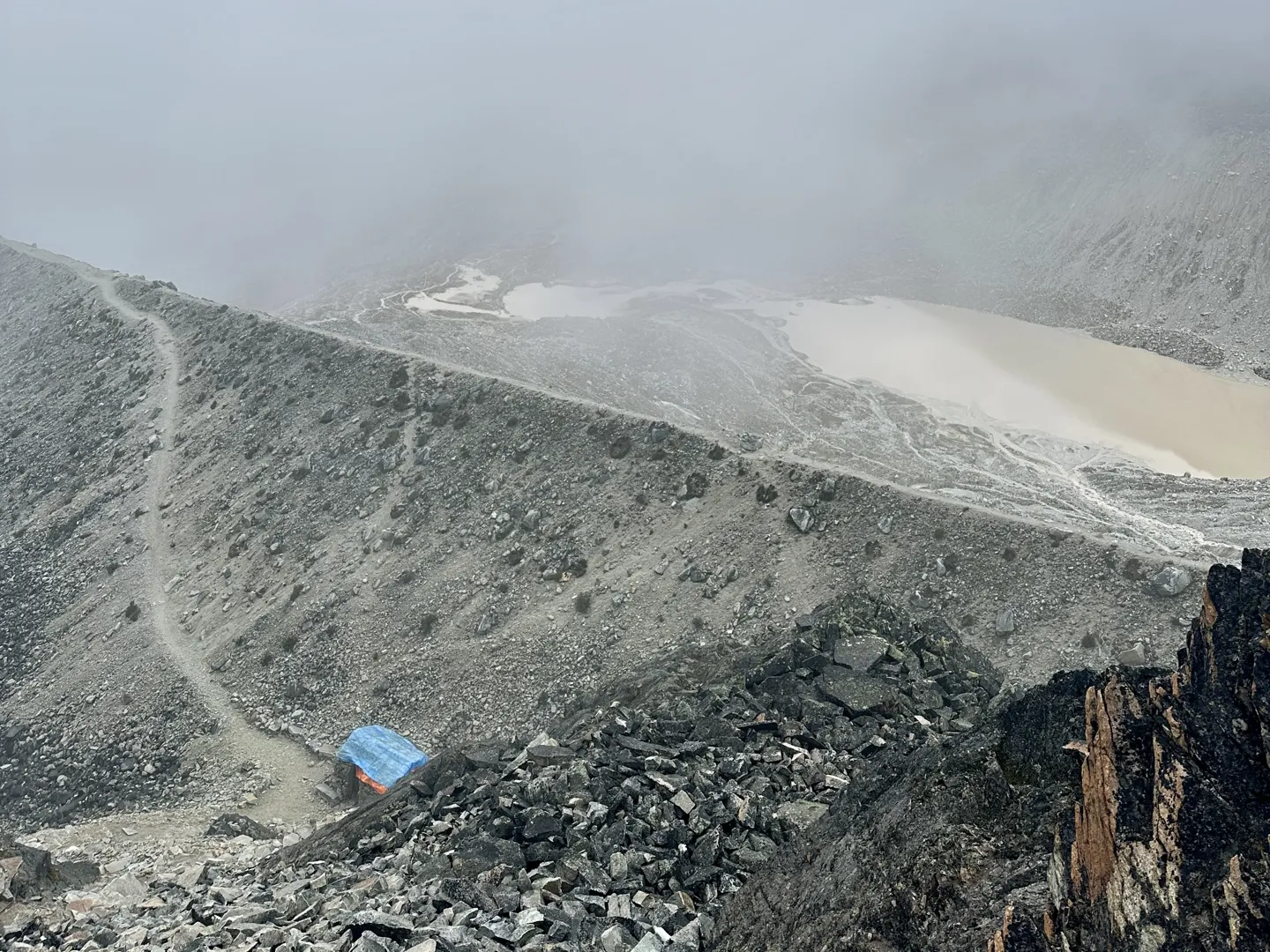 A small blue hut on the rocky moraine, cloud rolling across the pale glacial basin beyond