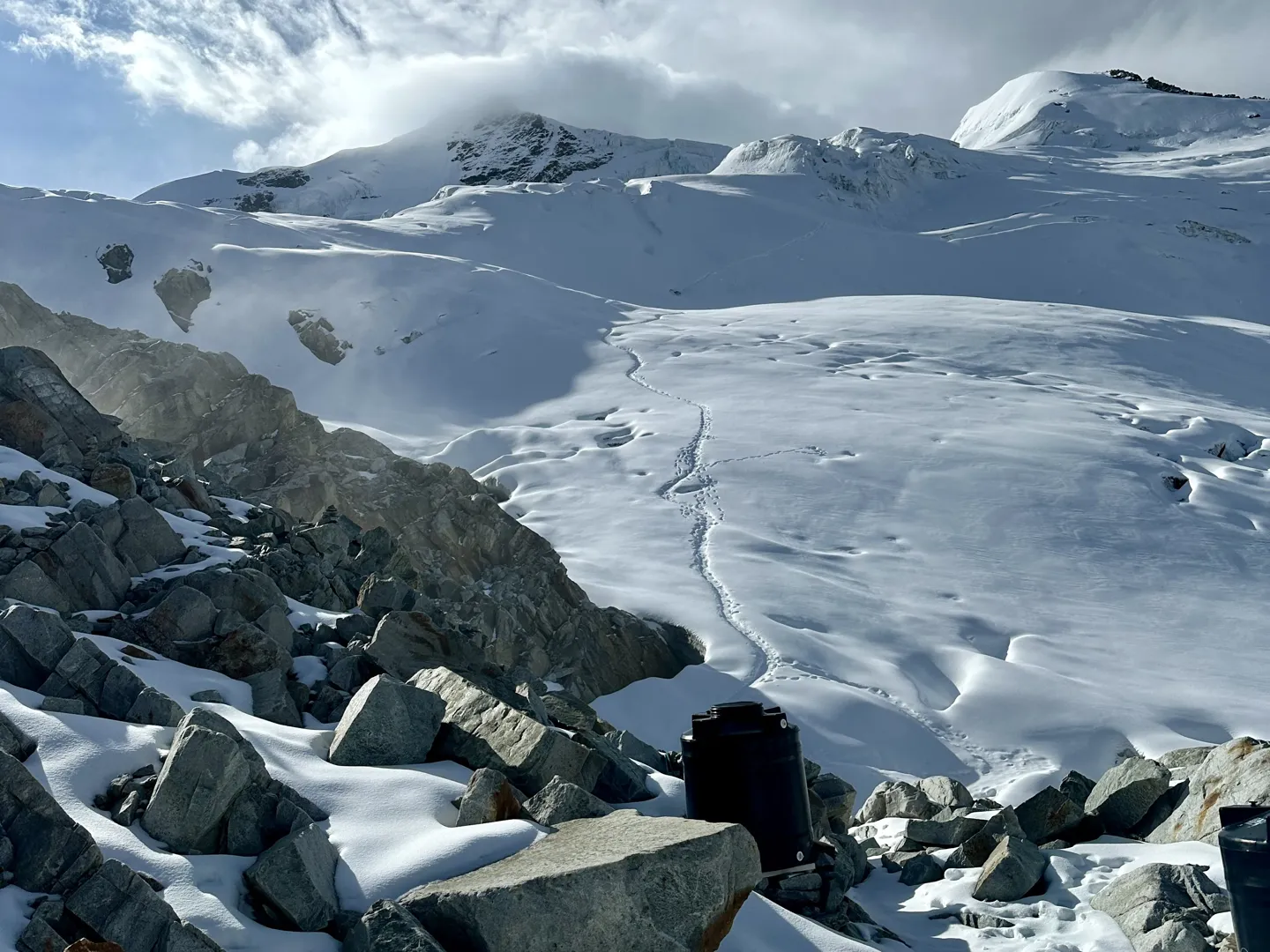 Boot tracks winding up the snow slopes above high camp, rocks and a water barrel in the foreground