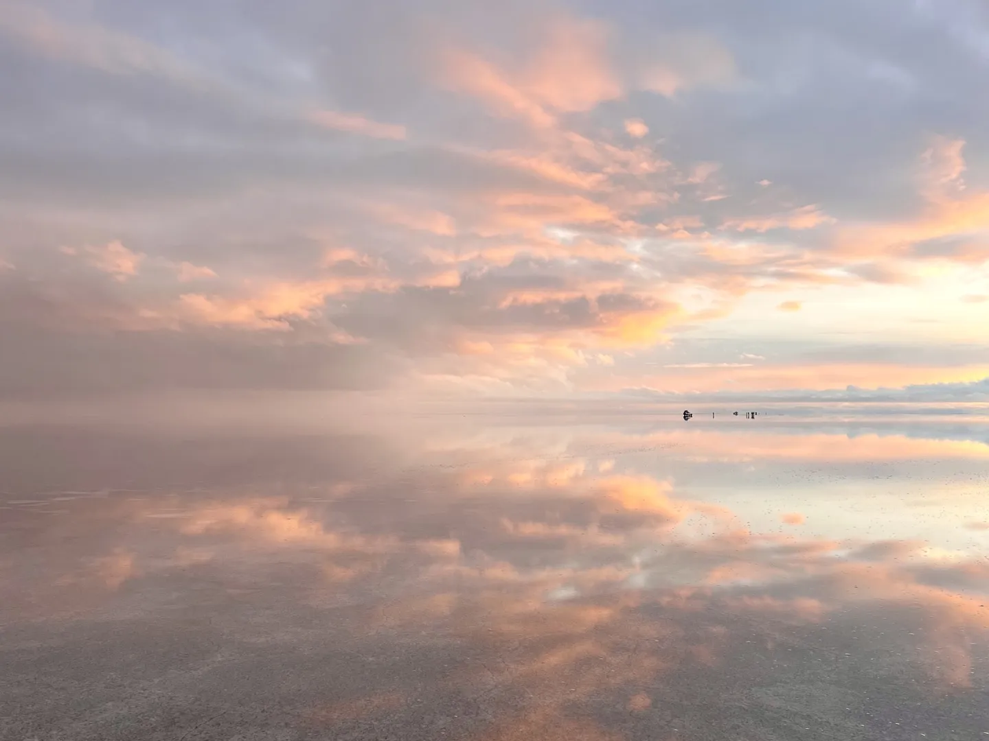 Pink sunrise clouds mirrored on the Salar de Uyuni with a 4WD and its passengers tiny in the distance