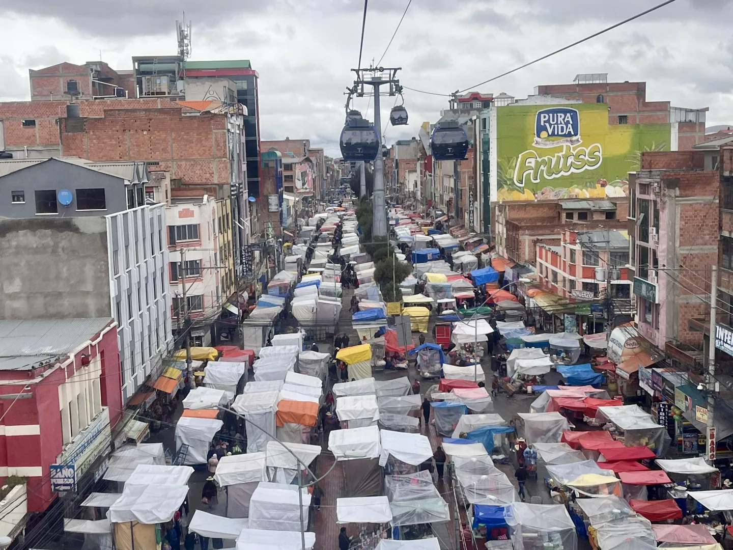 Rows of market stall canopies packed down a long La Paz street with cable cars crossing overhead