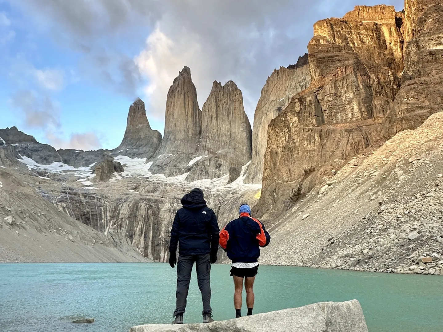 Two hikers standing on a rock by the lake, facing the towers at Mirador Base Torres at sunrise