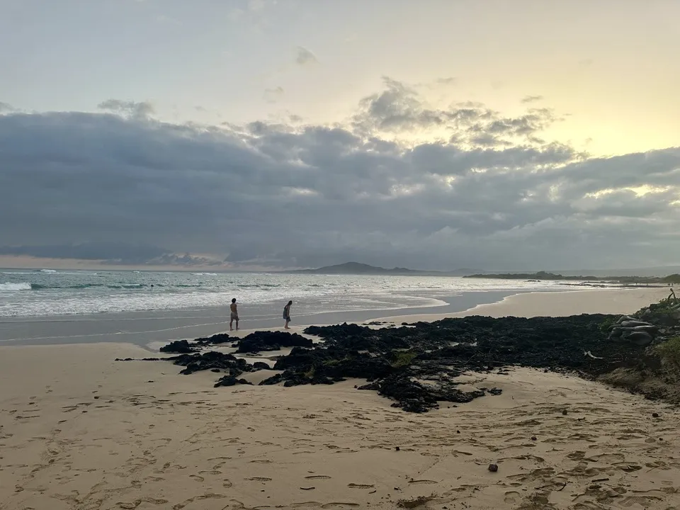 Two people at the water’s edge on a wide sandy beach at dusk, with black lava rocks in front