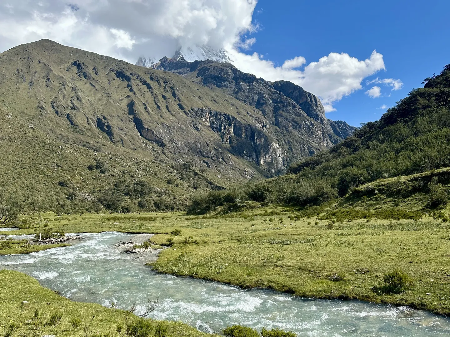 A milky glacial stream winding across a green valley floor, a snow-capped Cordillera Blanca summit rising into cloud