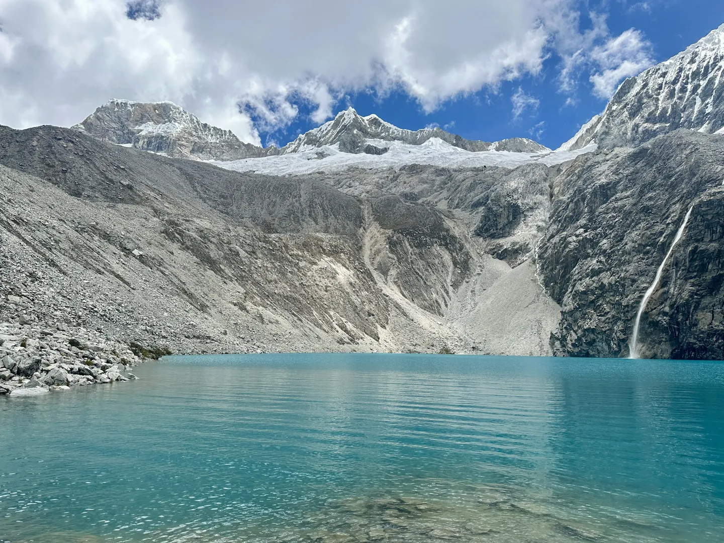 Laguna 69 seen from the shoreline, turquoise glacial water below scree slopes with a waterfall dropping from the snow-capped peaks