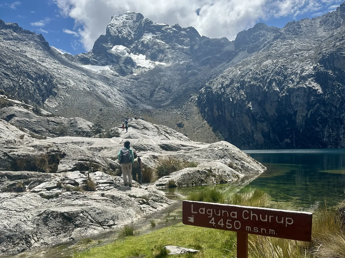 Hikers at the Laguna Churup sign marking 4,450 m, the lake and snow-capped Cordillera Blanca peaks behind