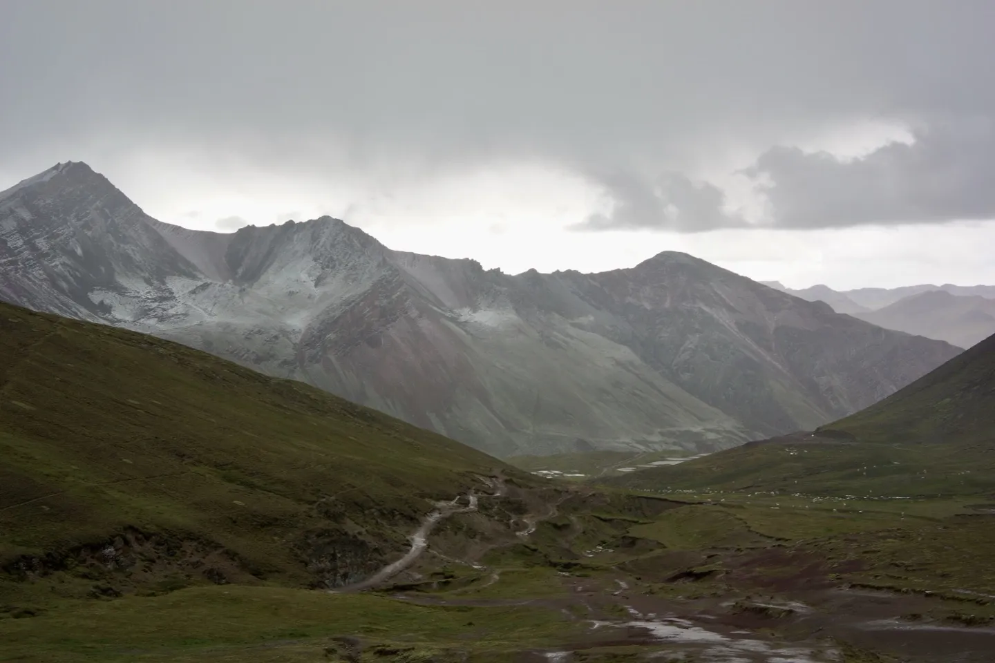 Snow-dusted Andean peaks above a dirt trail winding through a green valley near Rainbow Mountain